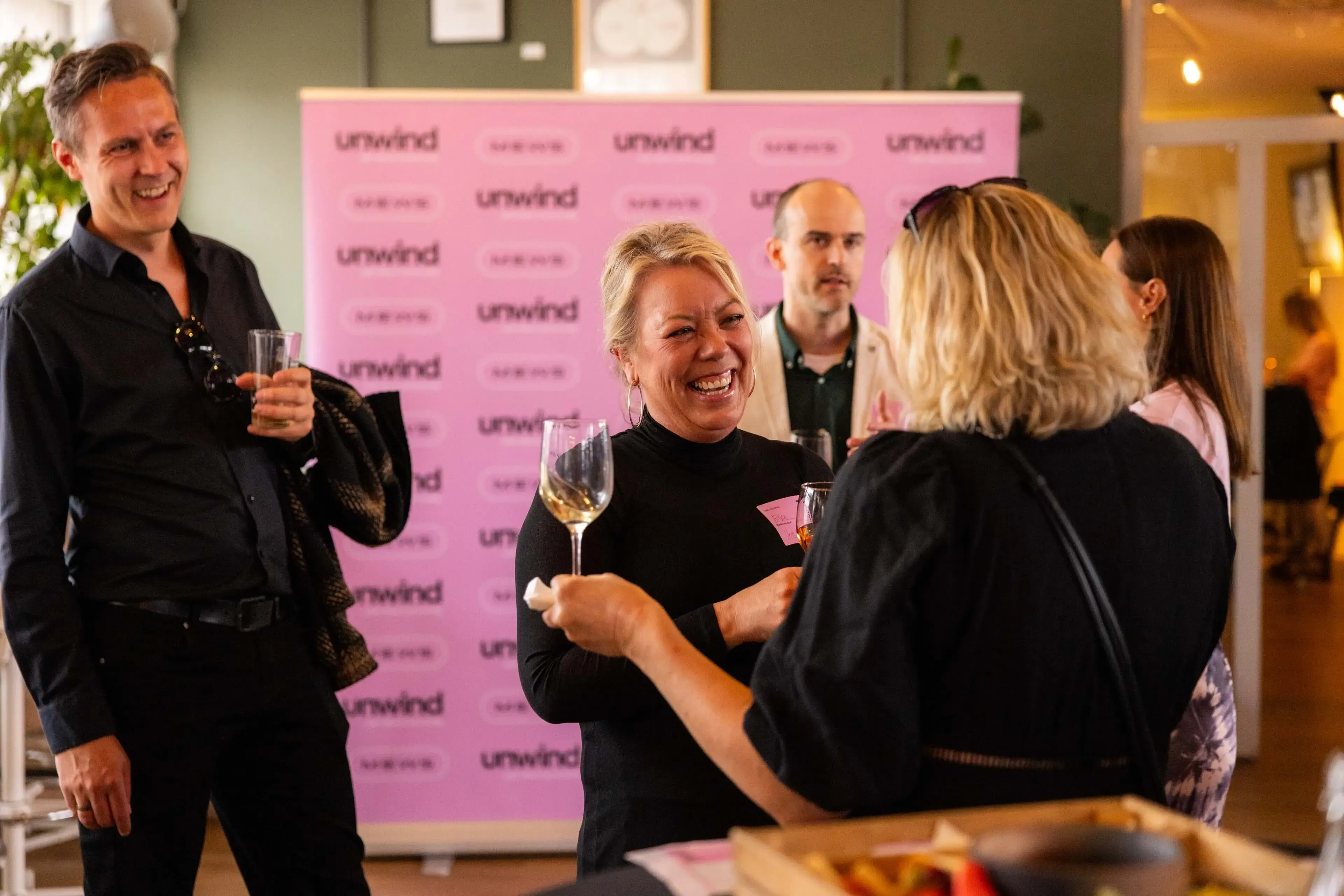 A group of people smiling and holding drinks while in conversation in front of a Mews Unwind banner.
