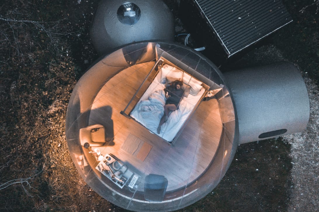 Woman lying on a bed inside a transparent, dome-shaped tiny house in a wooded area at dusk.