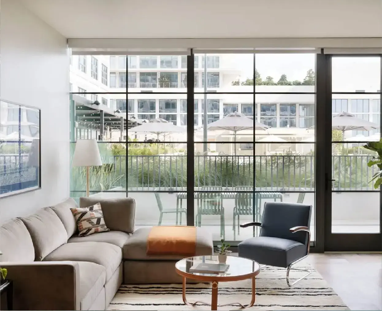 Wohnzimmer mit einem beigen Bereich, einem schwarzen Sessel, einem runden Couchtisch aus Holz und einem Balkon mit Stühlen und Sonnenschirmen im Freien.