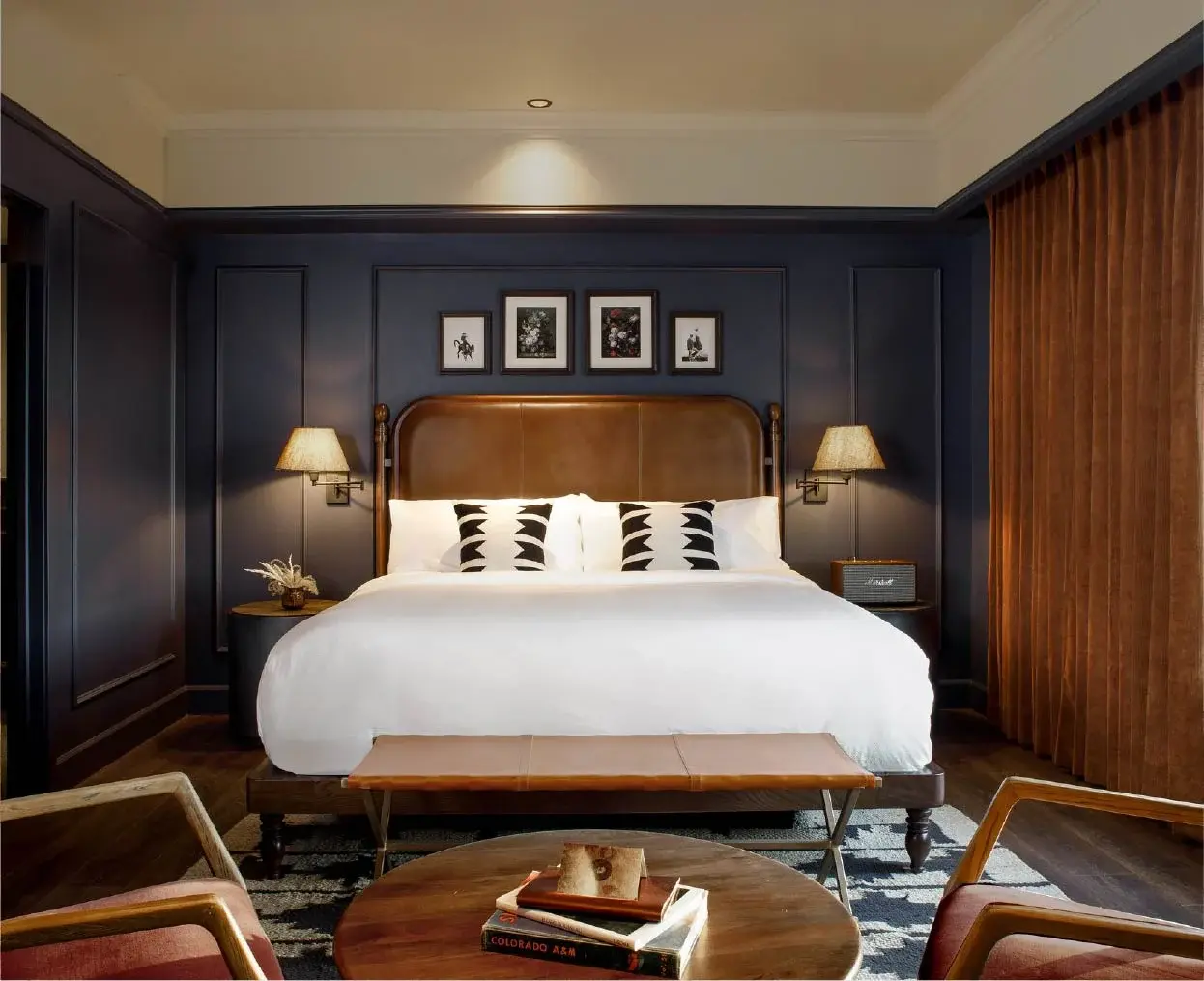 Een kingsize bed met wit beddengoed, zwarte en witte kussens, donkerhouten meubels en ingelijste kunst op een marineblauwe muur in een slaapkamer.