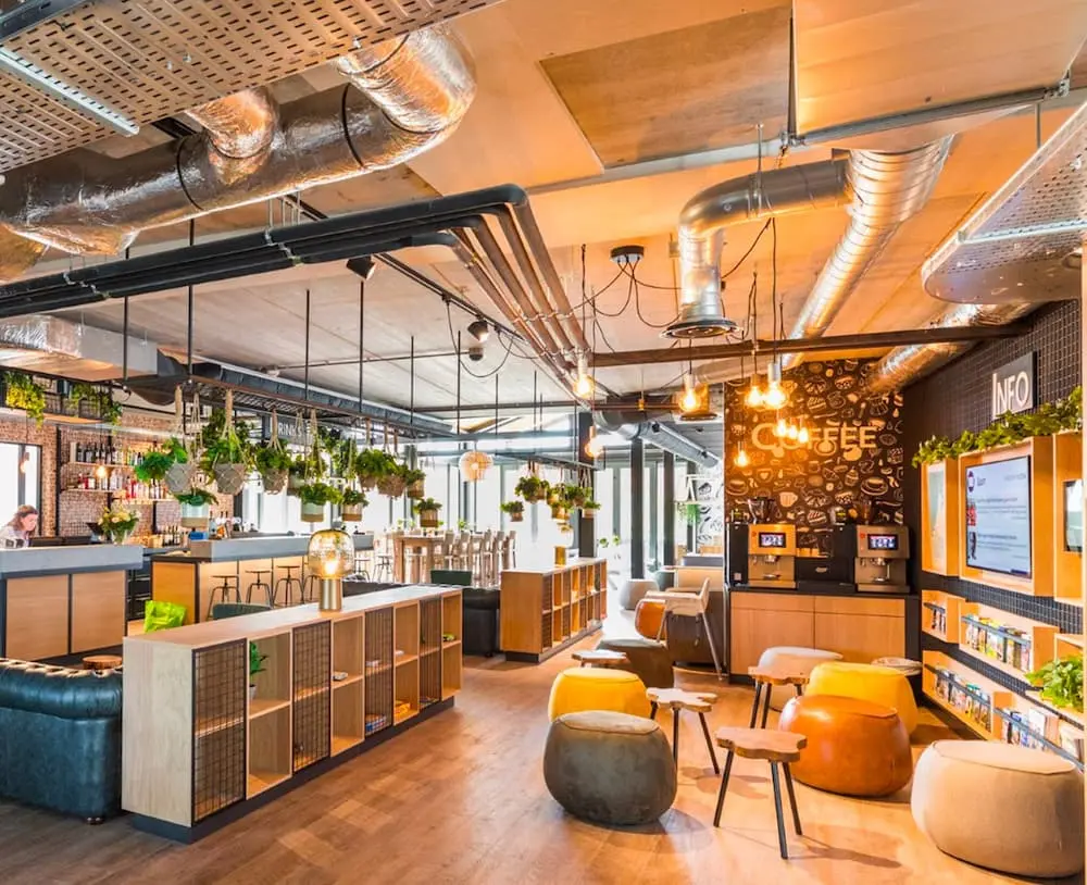 Moderne hotellobby met hangende planten, zitjes, houten accenten en zichtbare leidingen.