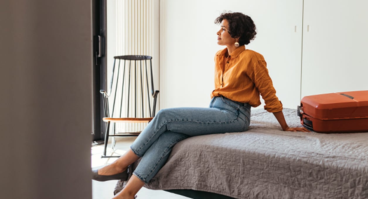 Vrouw zittend op een bed met een koffer naast zich, peinzend kijkend naar een raam of muur.