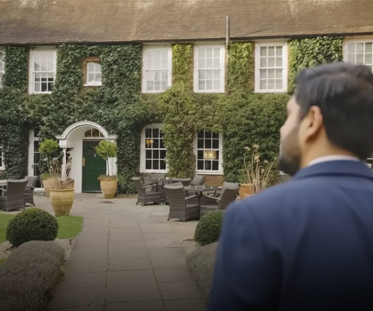 Man in business suit looking at a ivy-covered building with outdoor seating area.