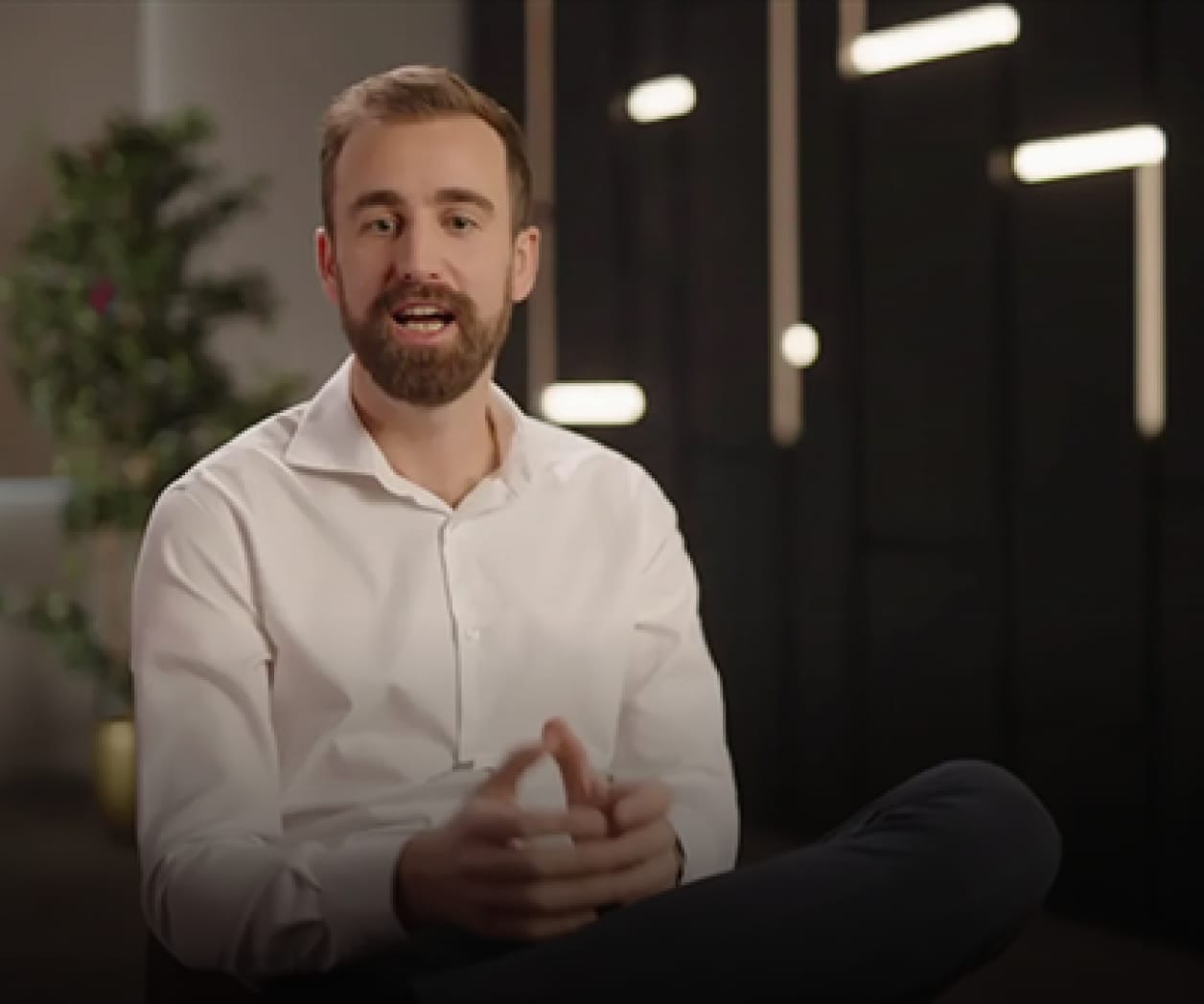 Bearded man in a white shirt speaking, seated with legs crossed in front of black panels with bright vertical lights.