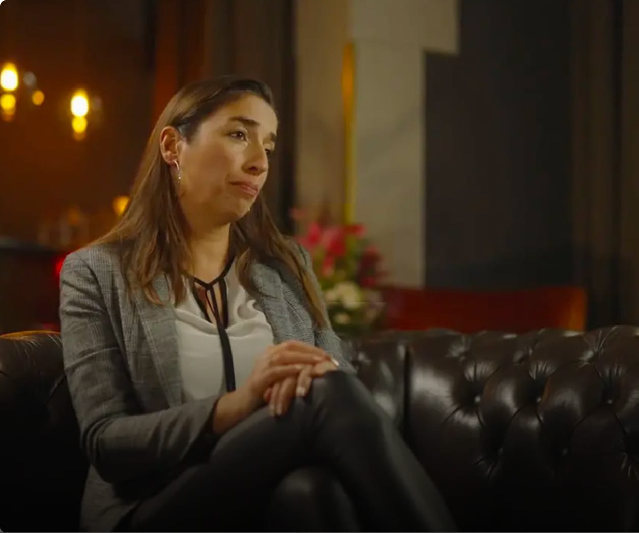 Woman sitting on a leather couch with a contemplative expression, wearing a gray blazer and white blouse, in a warmly lit room.