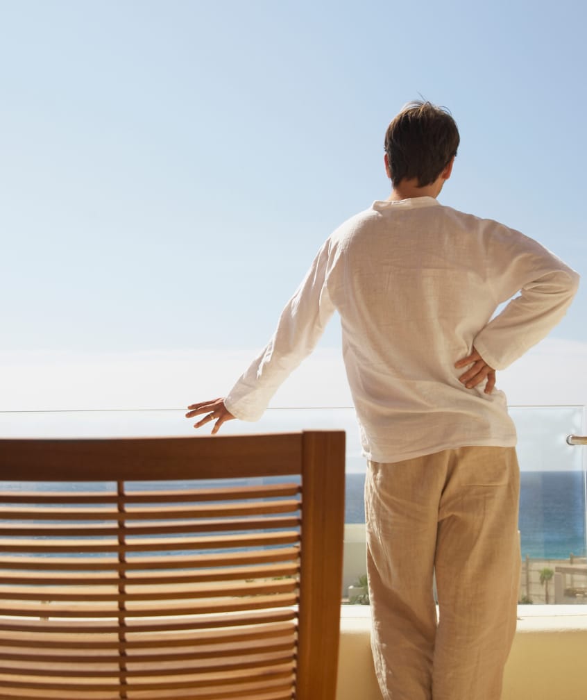 Person standing on a balcony looking at the ocean, with one hand on the railing and the other on their hip.