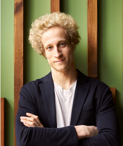 Jeune homme aux cheveux blonds bouclés, vêtu d'un blazer sombre et croisant les bras, debout sur un fond vert avec des panneaux verticaux en bois.