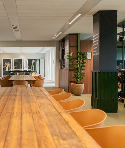 Langer Holztisch mit orangefarbenen Stühlen in einem modernen, minimalistischen Loungebereich mit einer hohen Topfpflanze und Akzenten in Schwarz und Holz.