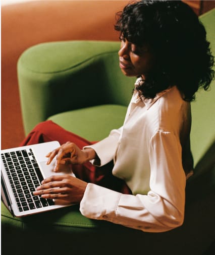 Woman with curly hair using a laptop while seated on a green armchair.