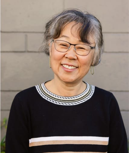 Femme âgée souriante aux cheveux gris courts et aux lunettes, portant un pull noir avec des détails blancs, debout contre un mur gris.