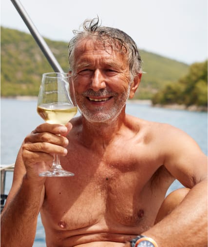 A smiling older man with gray hair and beard, shirtless, holding a glass of white wine outdoors near a body of water.