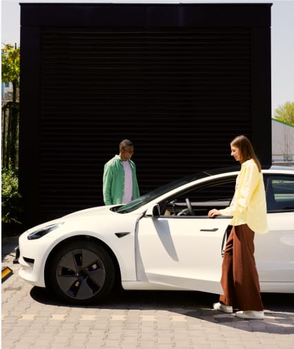 Two women are standing next to a white electric car parked in front of a black garage door.