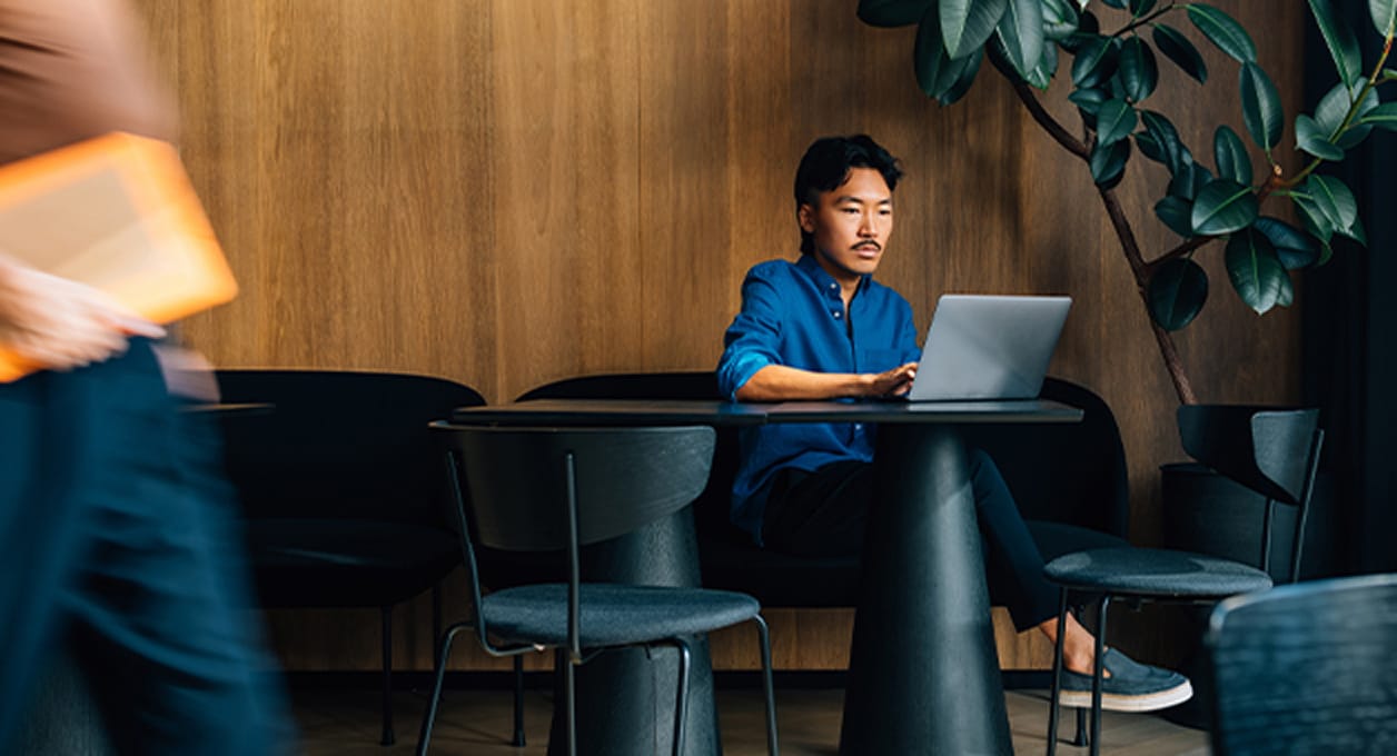 Man in een blauw shirt werkt op een laptop aan een zwarte tafel in een moderne, gezellige binneninstelling met houten muren en groene planten.