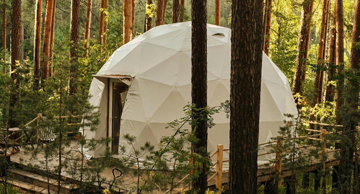 Geodetische koepeltent omringd door bomen in een bos met een houten dek en hekwerk.