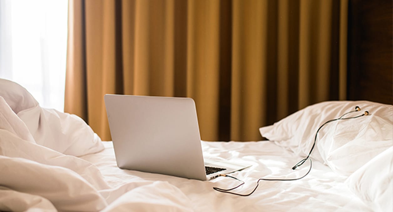 Laptop met koptelefoon rustend op verfomfaaid wit beddengoed in een slaapkamer.