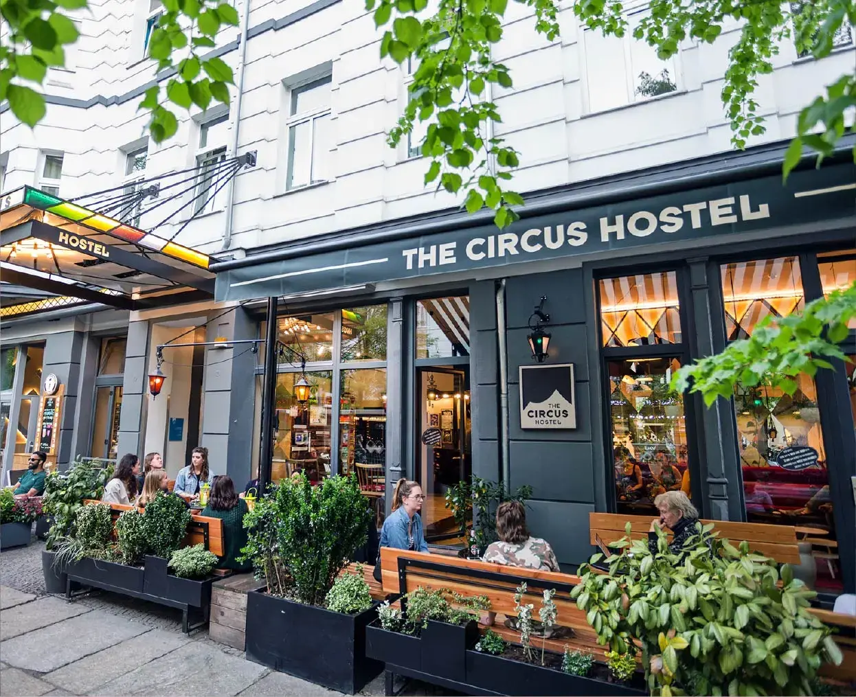 People sitting and chatting outside The Circus Hostel on a city street with green foliage overhead.