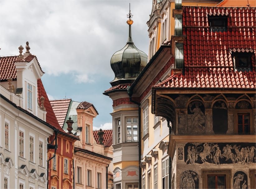 Colorful historic buildings with ornate facades and red tiled roofs in Vinohrady, Prague, under a partly cloudy sky.