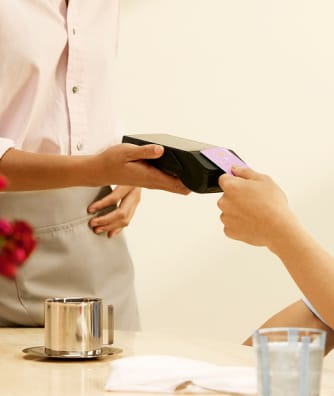 Person's hand holding a payment terminal, receiving a card from another person, at a table with a cup and a glass.